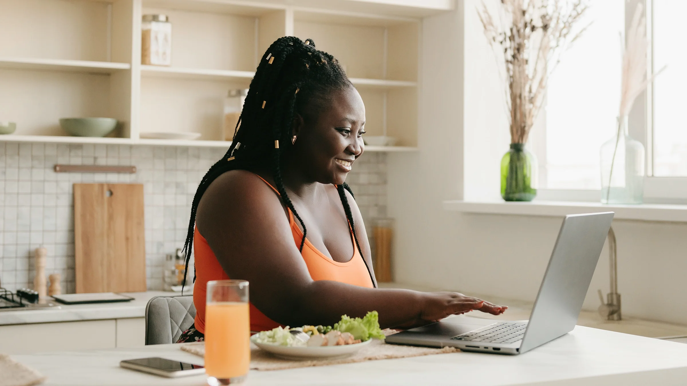 A woman has a nutritious lunch of veggies and juice while working on her laptop. 