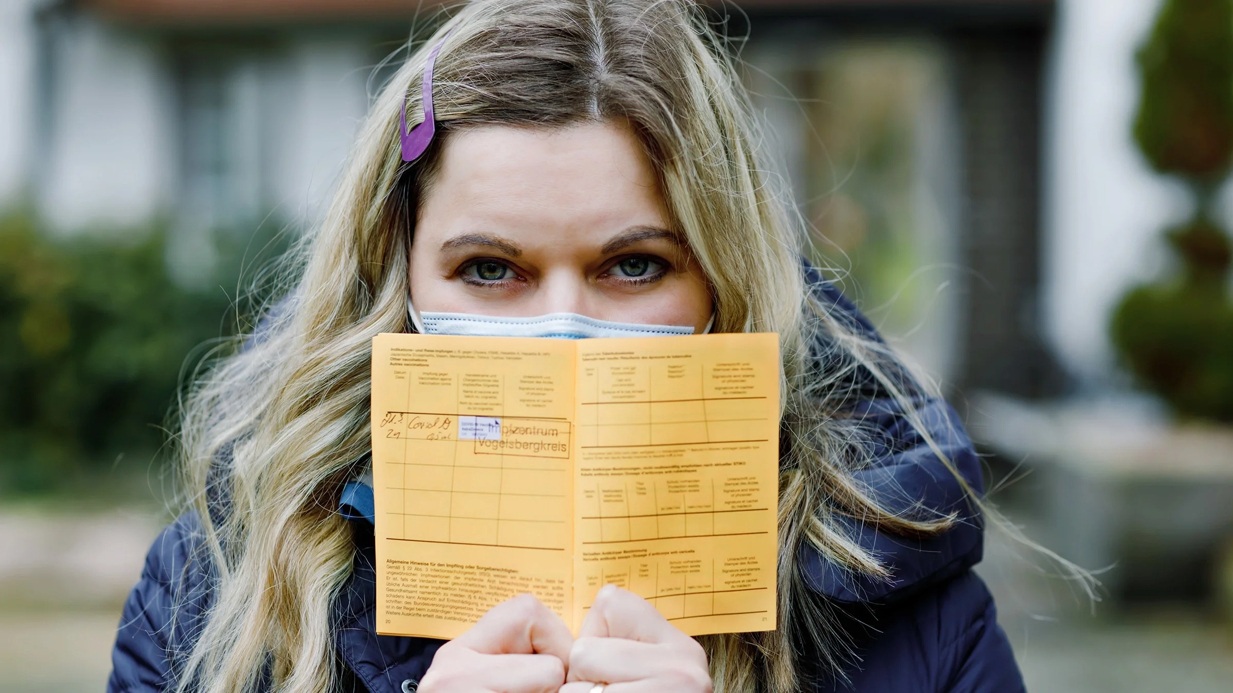 Portrait of a woman holding up an orange vaccine passport in front of her face. She is wearing a blue medical face mask and a puffy blue raincoat.