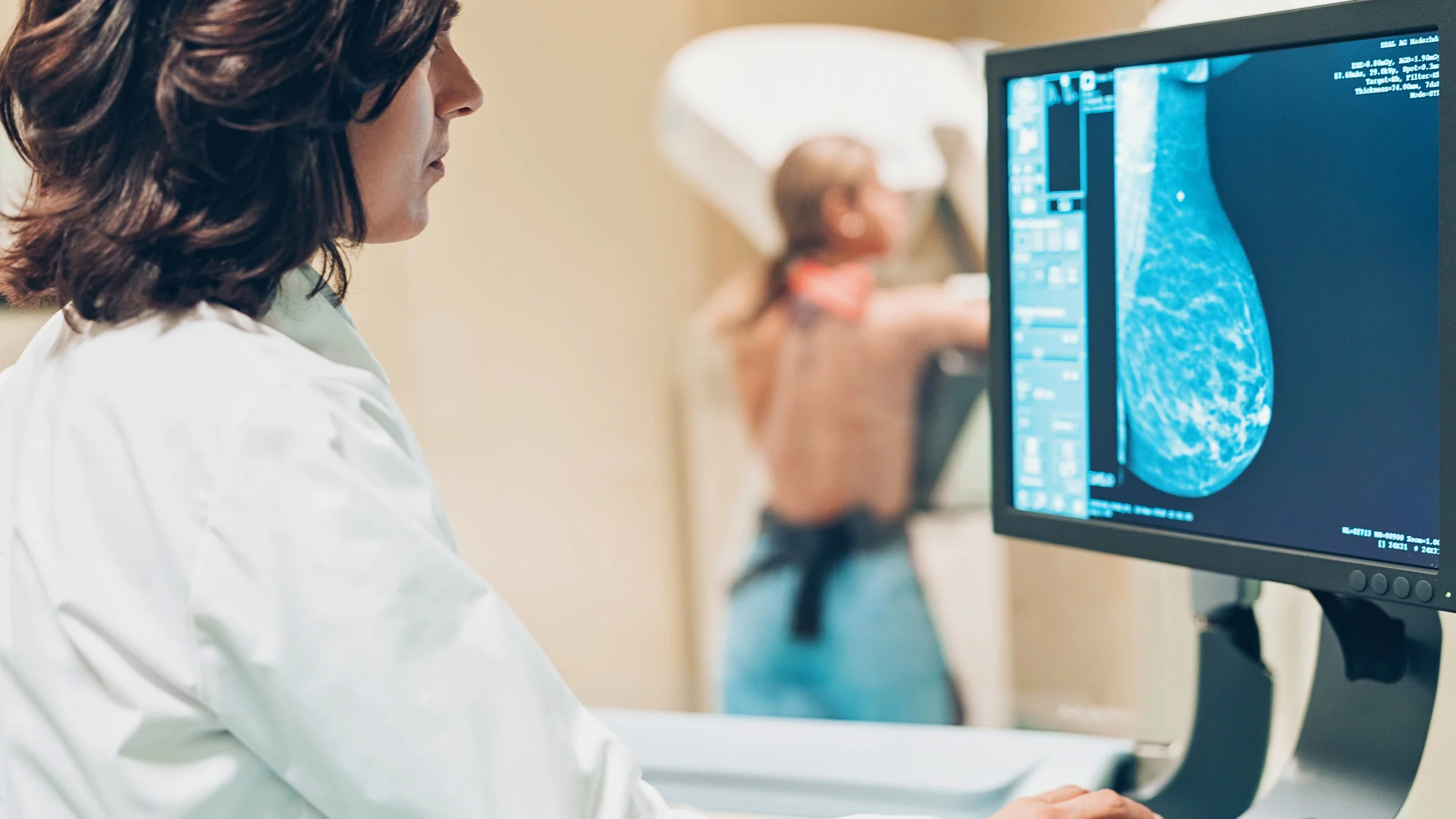 A doctor examining a breast mammogram on their screen. The patient is in the background.