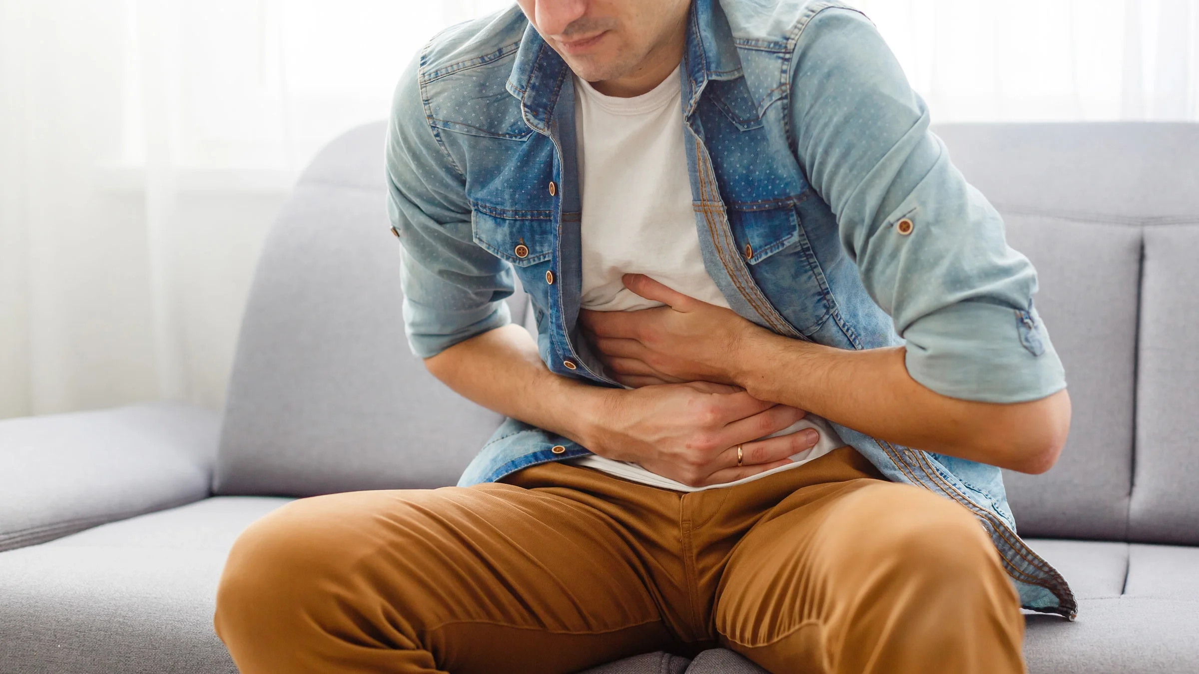 A man sitting on a gray couch holding his stomach in pain.
