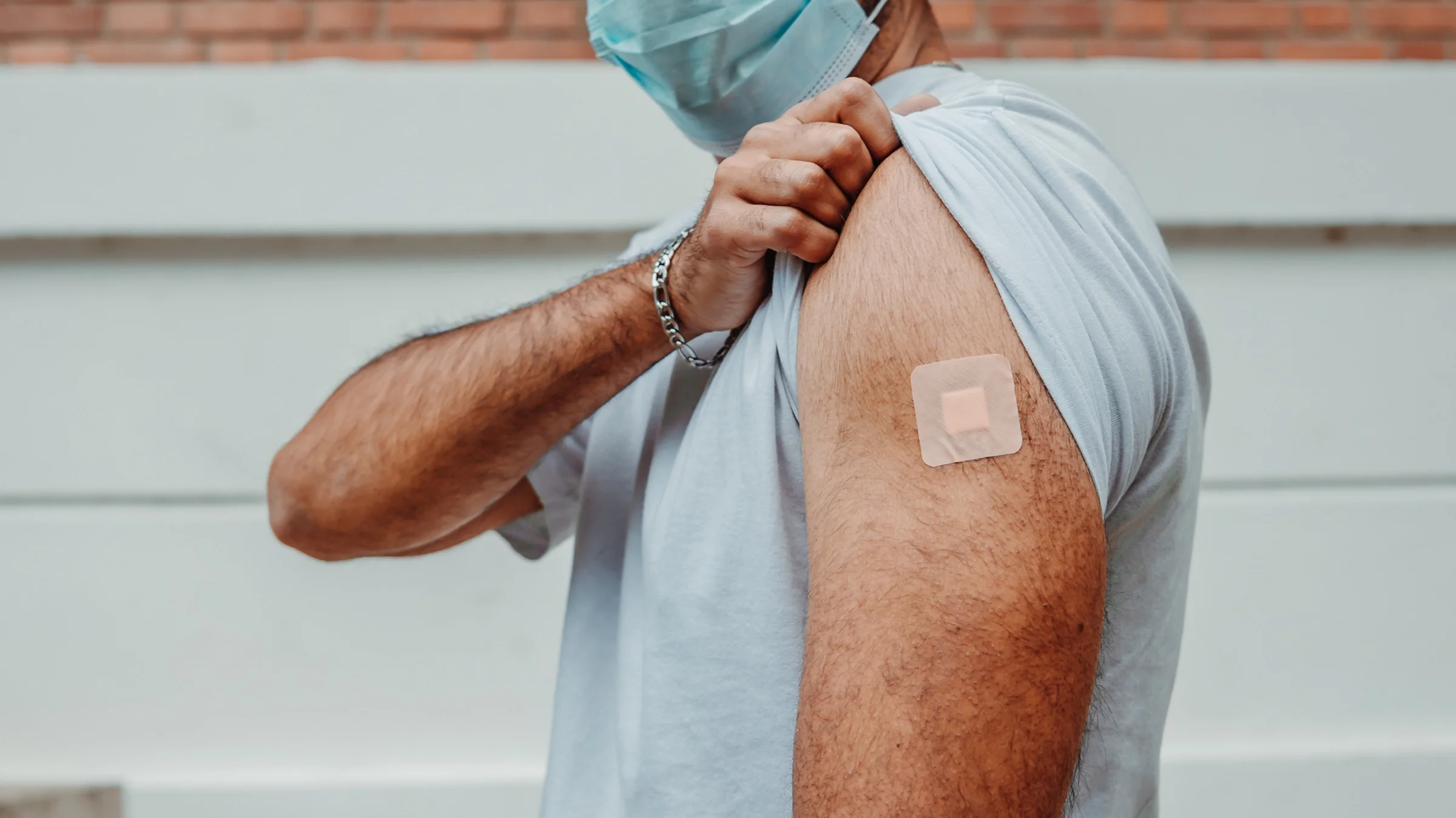 Young man wearing a blue medical face mask pulling up his sleeve to show his band-aid from the COVID-19 vaccine.