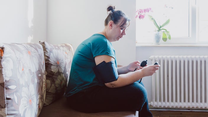 A woman measures her blood pressure with a machine while sitting on a couch.