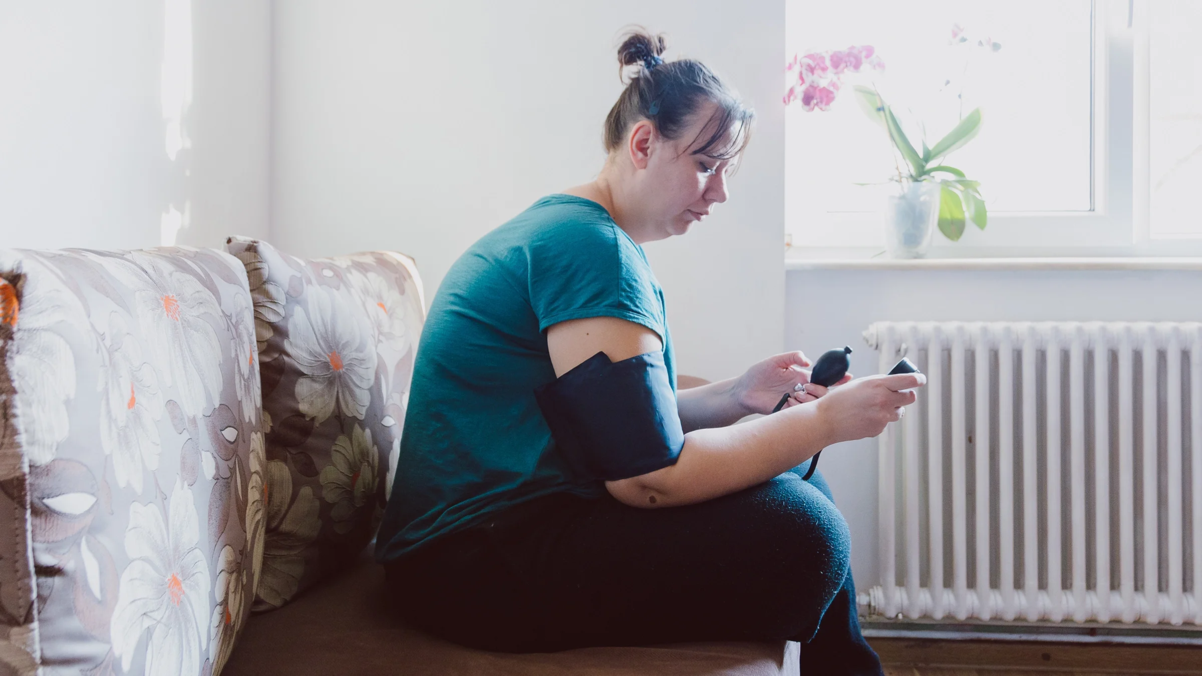 A woman measures her blood pressure with a machine while sitting on a couch.