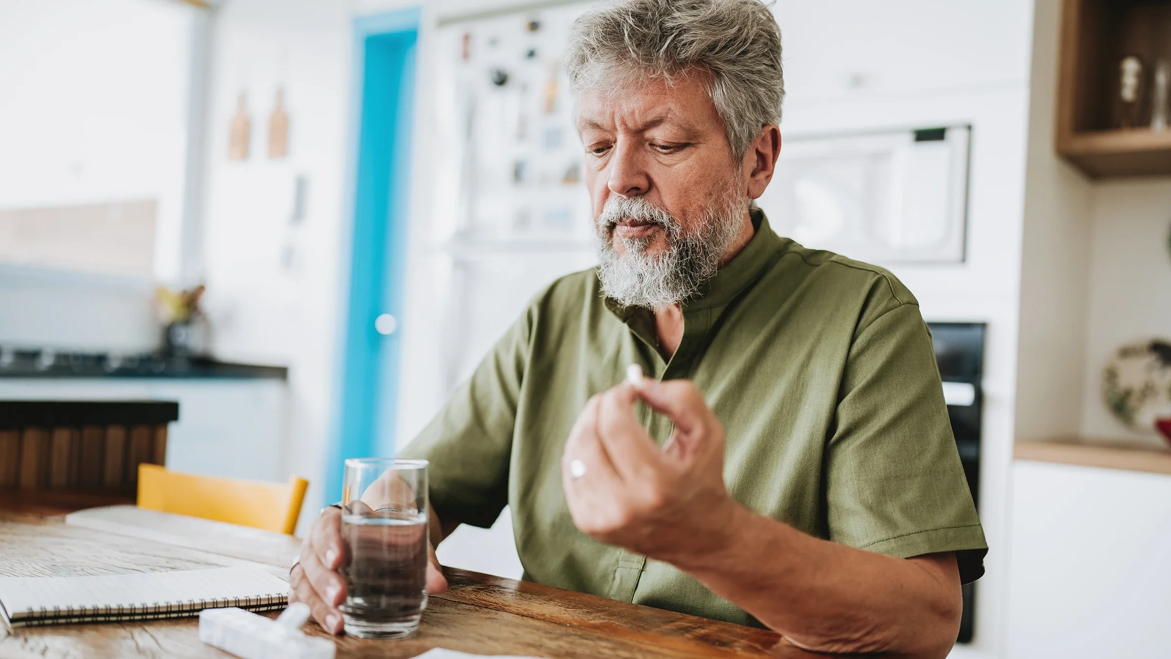 A man is taking medication with a glass of water at the kitchen table. 