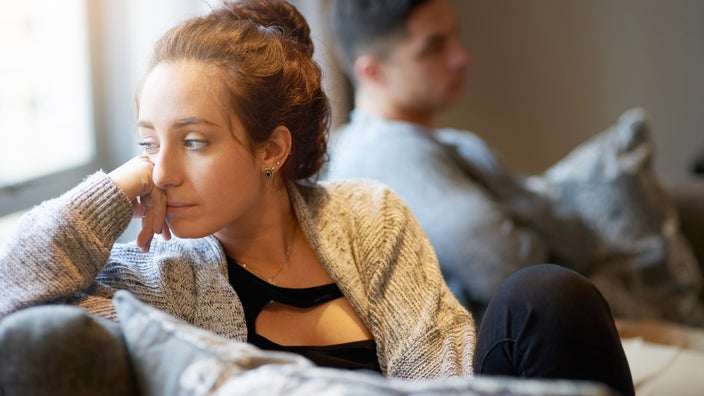 A person thinking on their couch after an argument. Their partner is in the background.