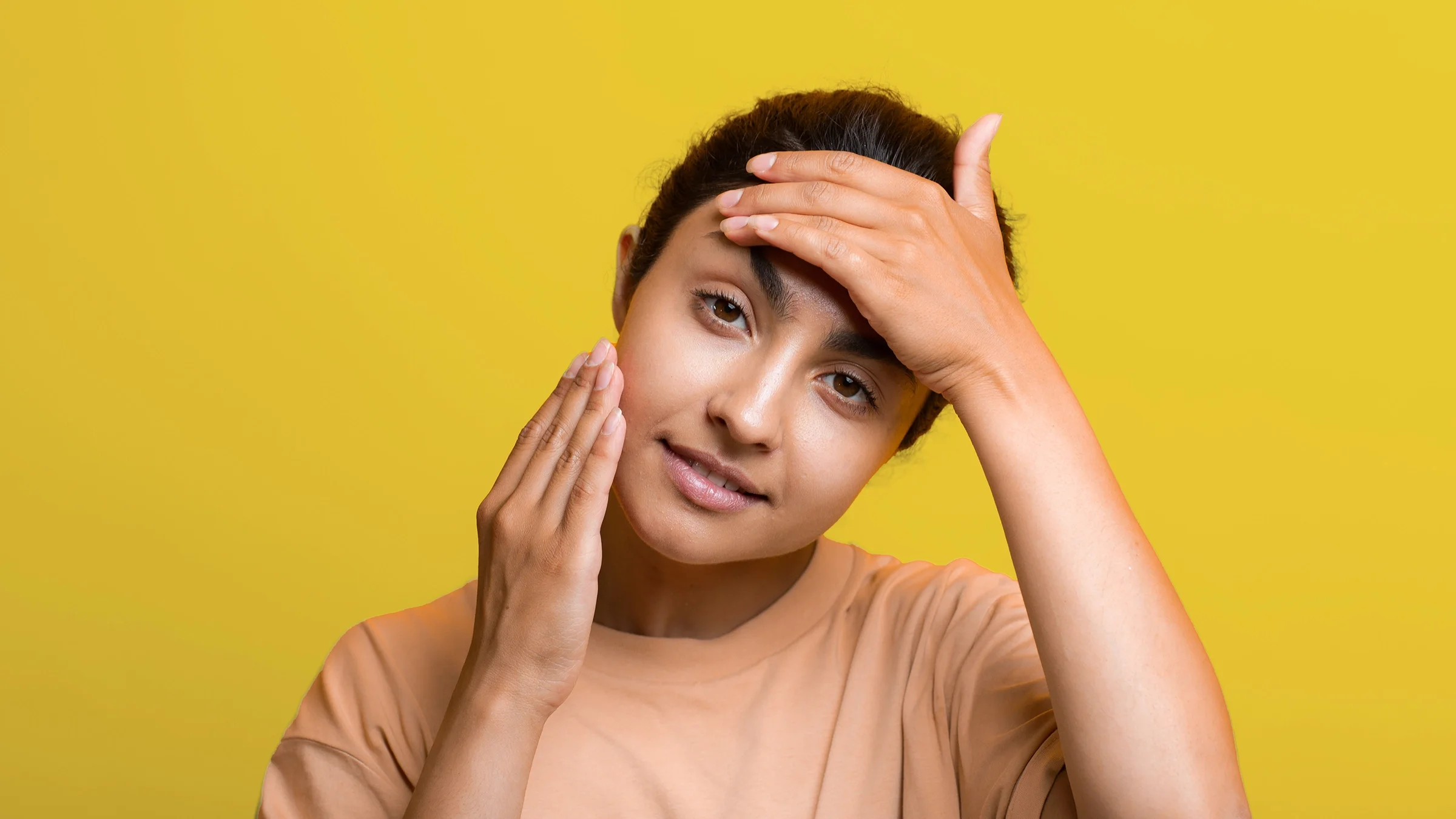 Portrait of a young woman doing face yoga on a plain yellow studio background.