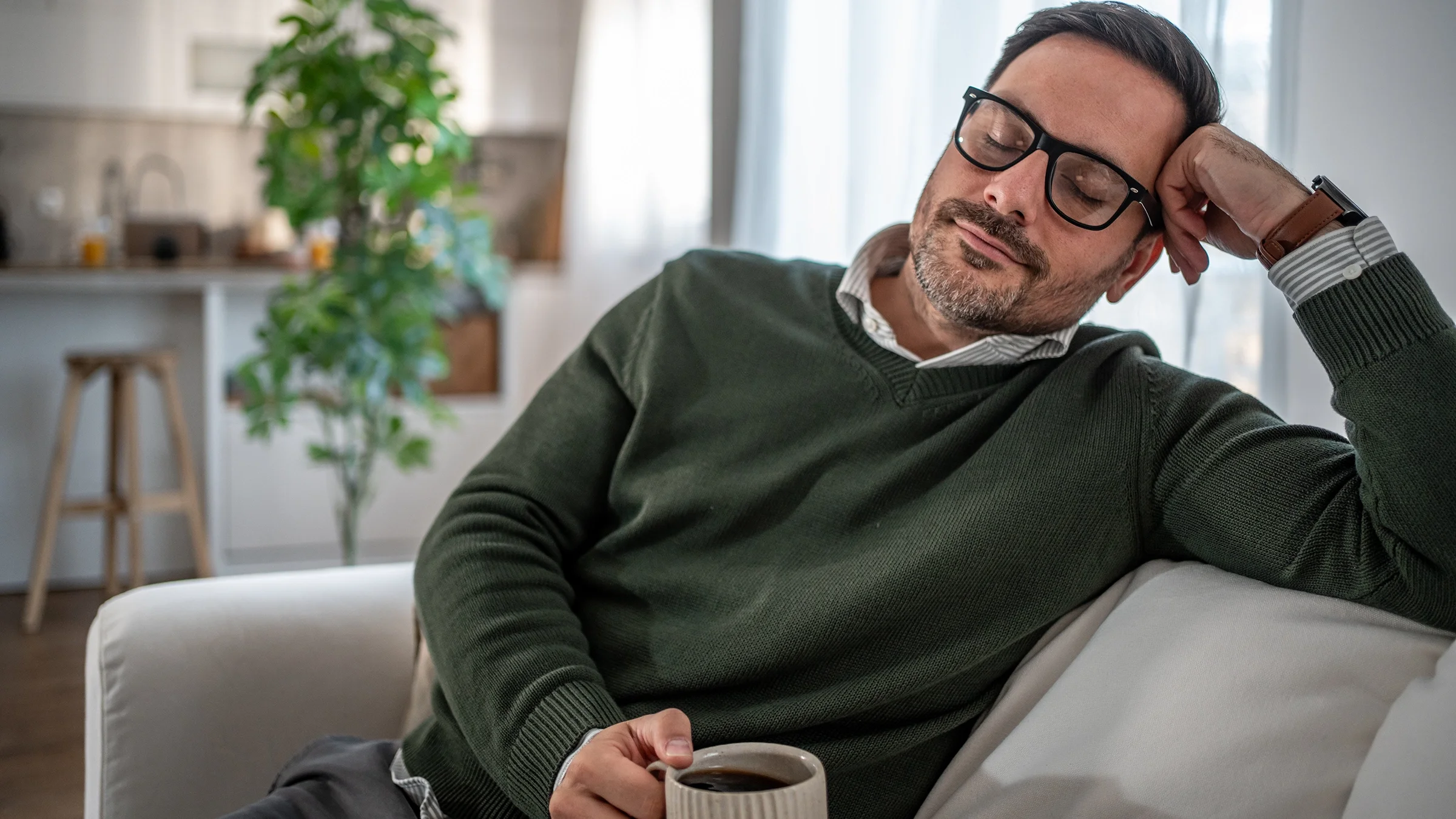 Man napping on sofa in living room holding coffee.