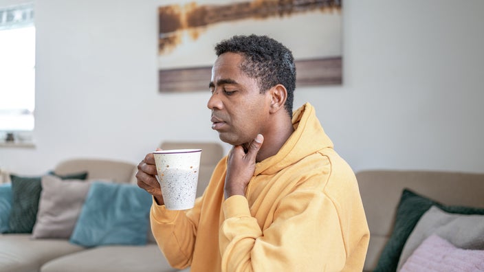A man is at home rubbing his sore throat while holding a cup of tea.