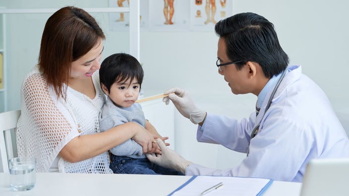A pediatrician asking a young child who is sitting on their parent's lap to open their mouth to examine it with a tongue depressor.
