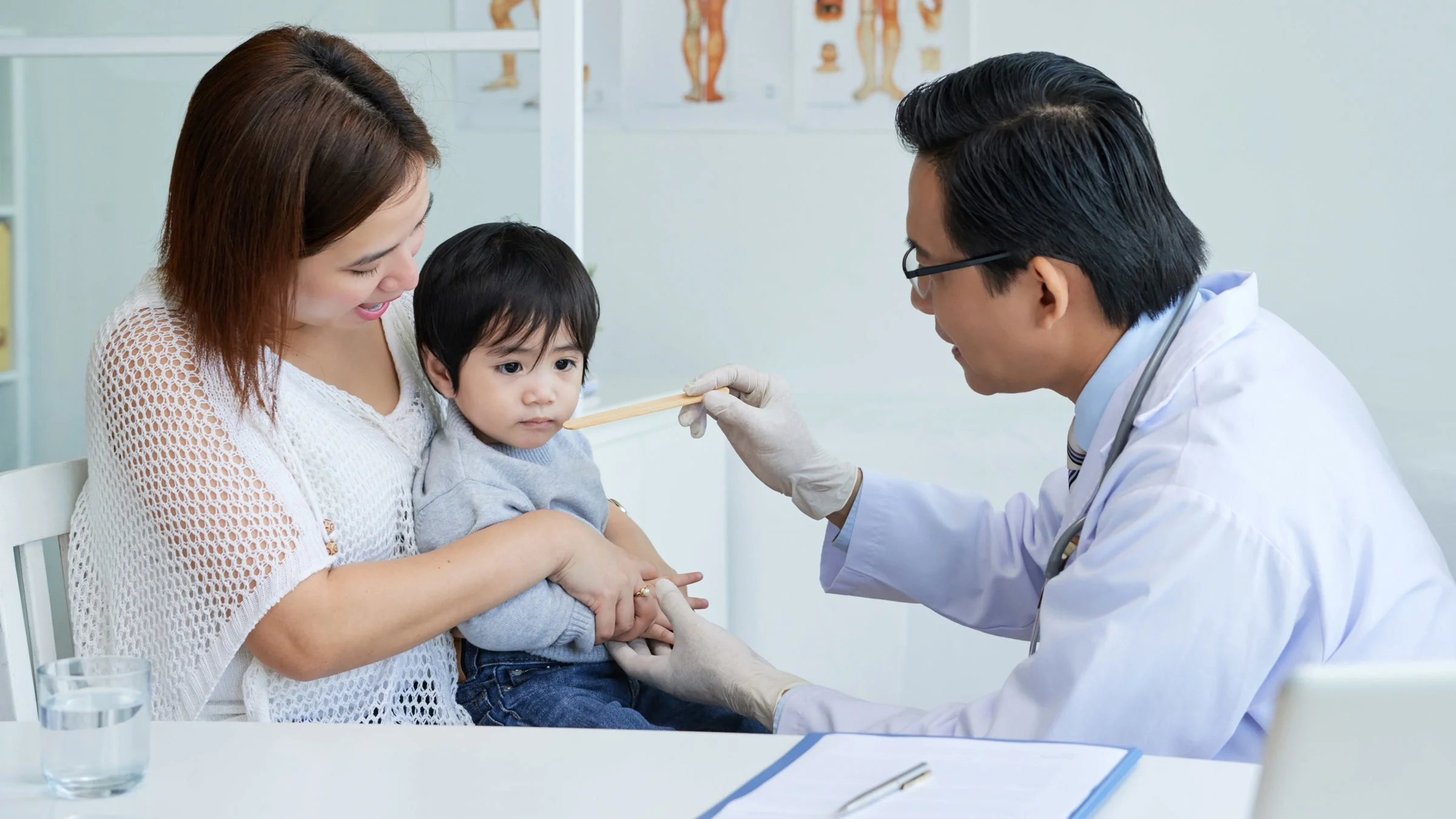 A pediatrician asking a young child who is sitting on their parent's lap to open their mouth to examine it with a tongue depressor.