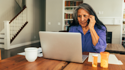 A woman is talking on the phone while looking at her laptop, which is next to bottles of medication on her kitchen table.
RichVintage/E+ via Getty Images