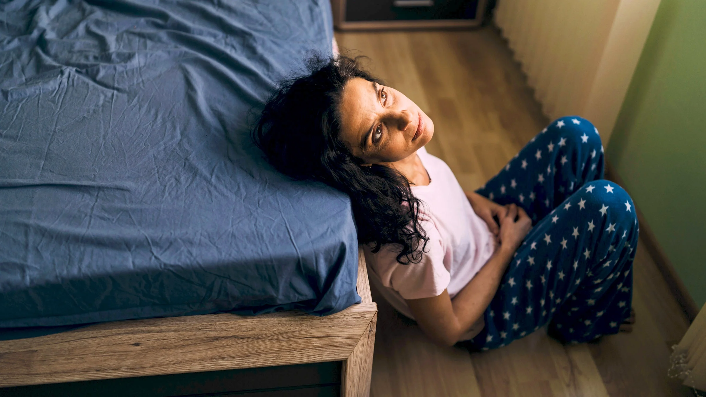 A sad middle-aged person by the foot of their bed.