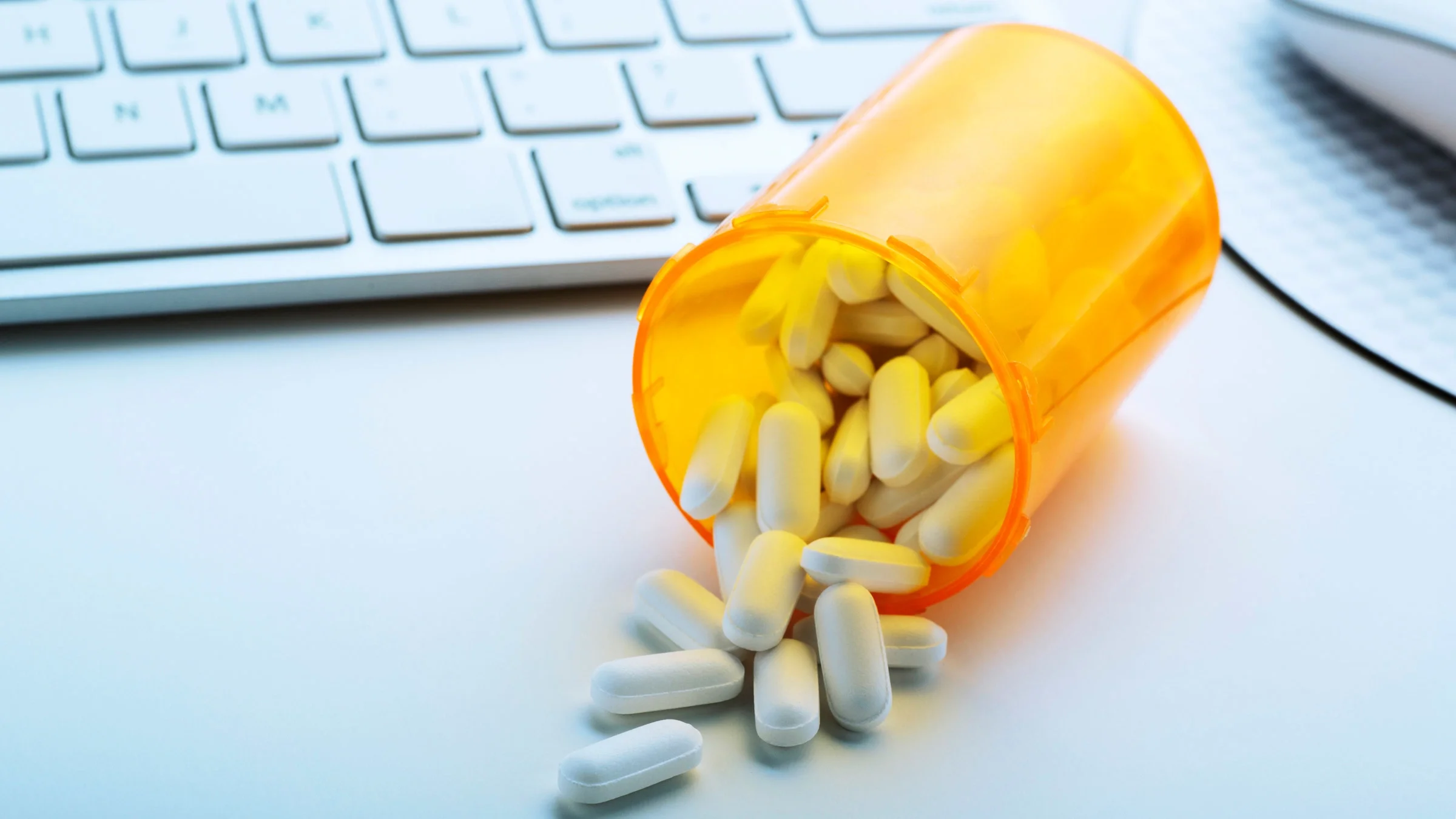 Pills spilling out of a bottle onto a desk, next to a keyboard and mouse.