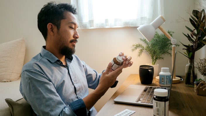 Young man reading the back of the medication bottle. He is sitting at his desk at home.
