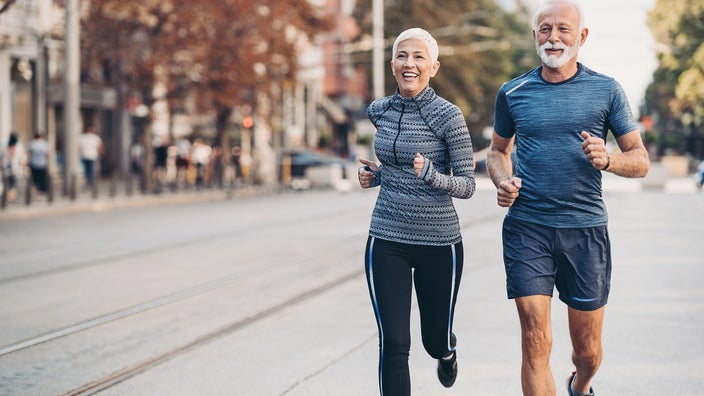Senior couple on a run through the city. Both have white hair and are smiling.