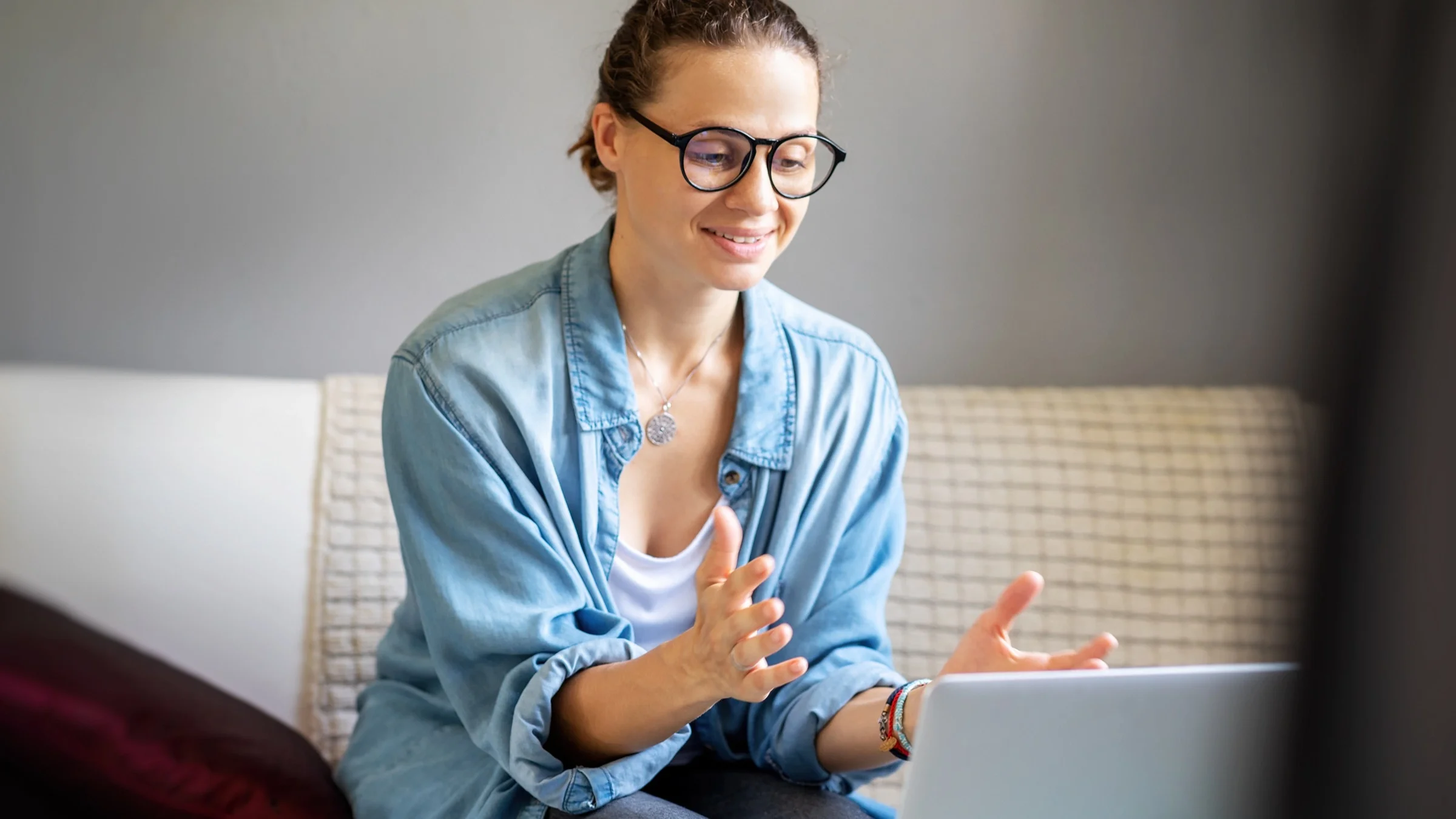 A young woman sitting on her couch using her laptop for a telehealth appointment. She has her hand out and palm up like she is explaining something.
