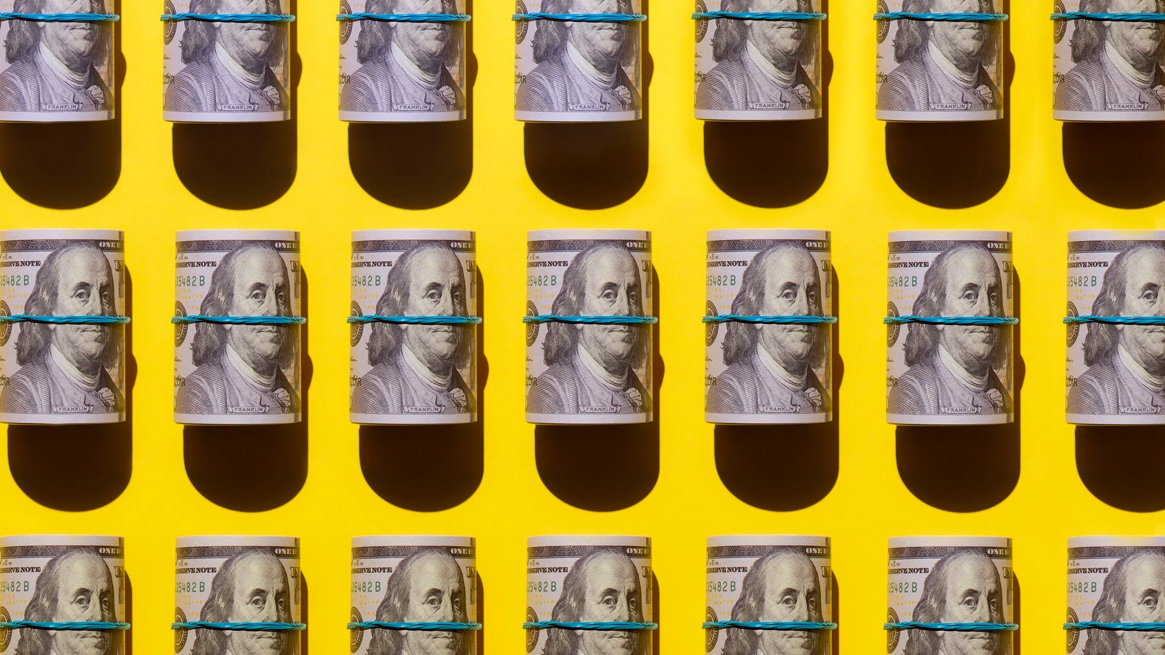 Rubber banded rolls of cash lined up in rows across the frame. There are three rows in total from top to bottom. They are on a bright yellow background.