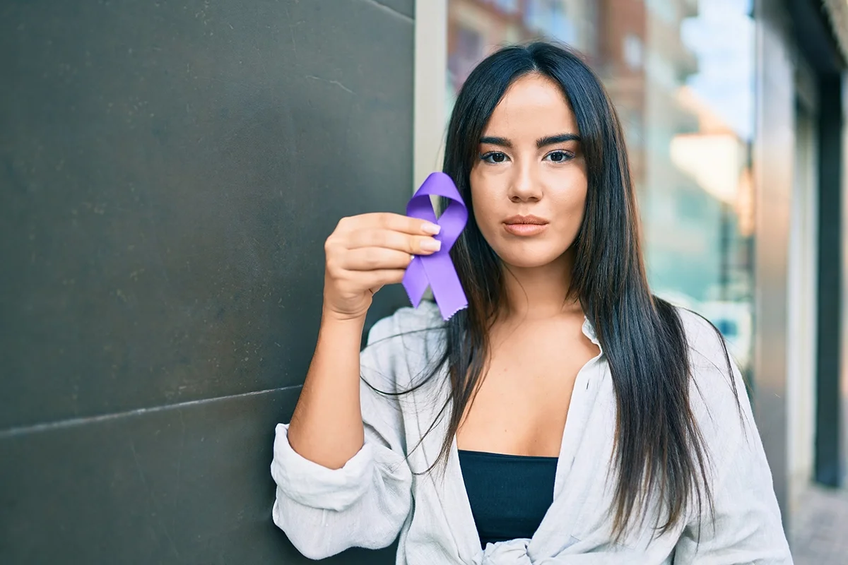 Portrait of a young woman holding up a purple ribbon standing outside.