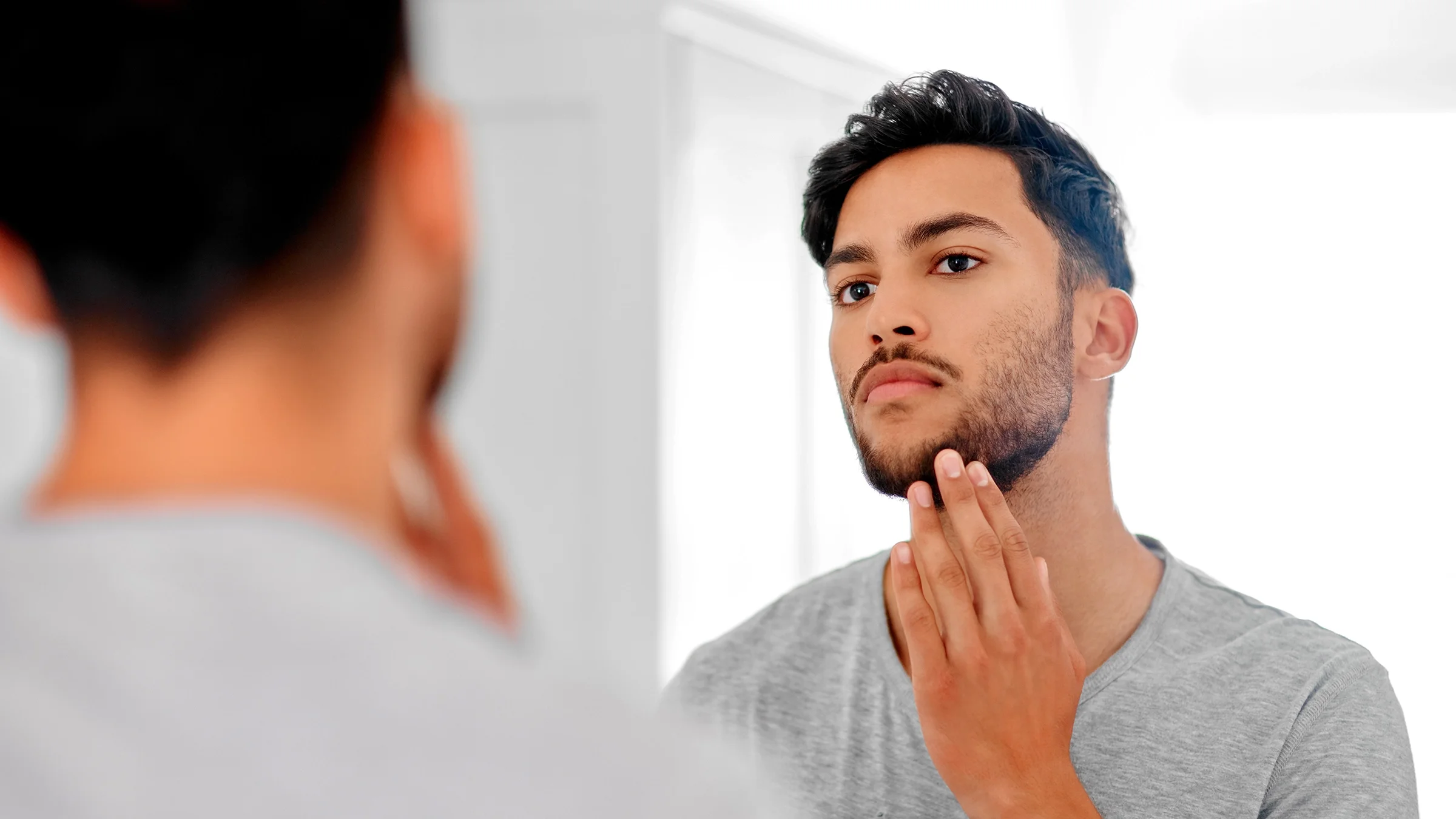 A man is looking at his beard in the mirror at home.