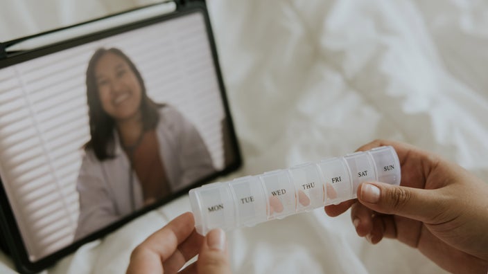 A person's hands holding a pill container. There's a pharmacist on a tablet screen in the background.