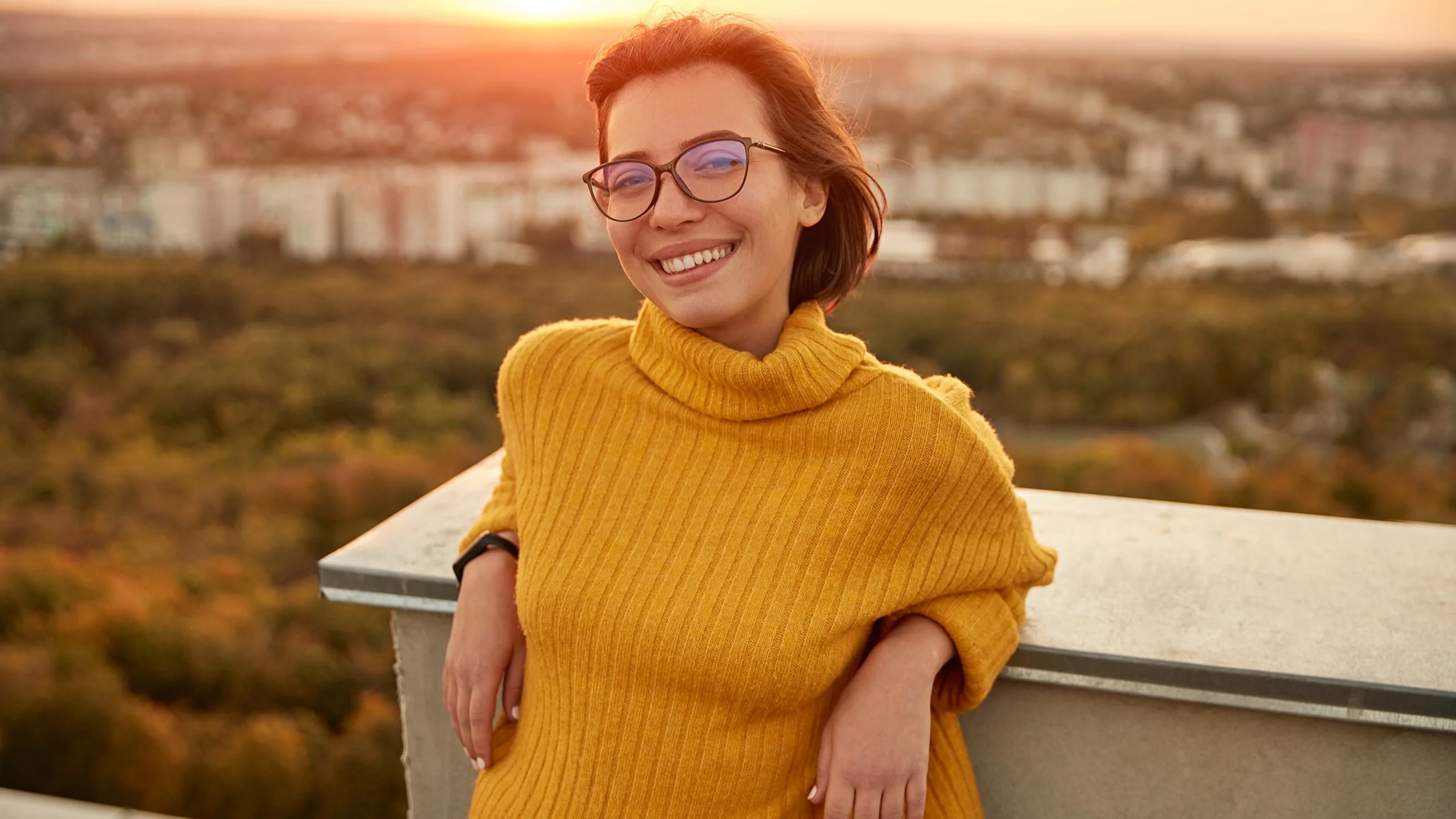 A portrait of a young woman with short hair and glasses. She is leaning back on a ledge smiling. There is a beautiful golden-pink sunset behind her.