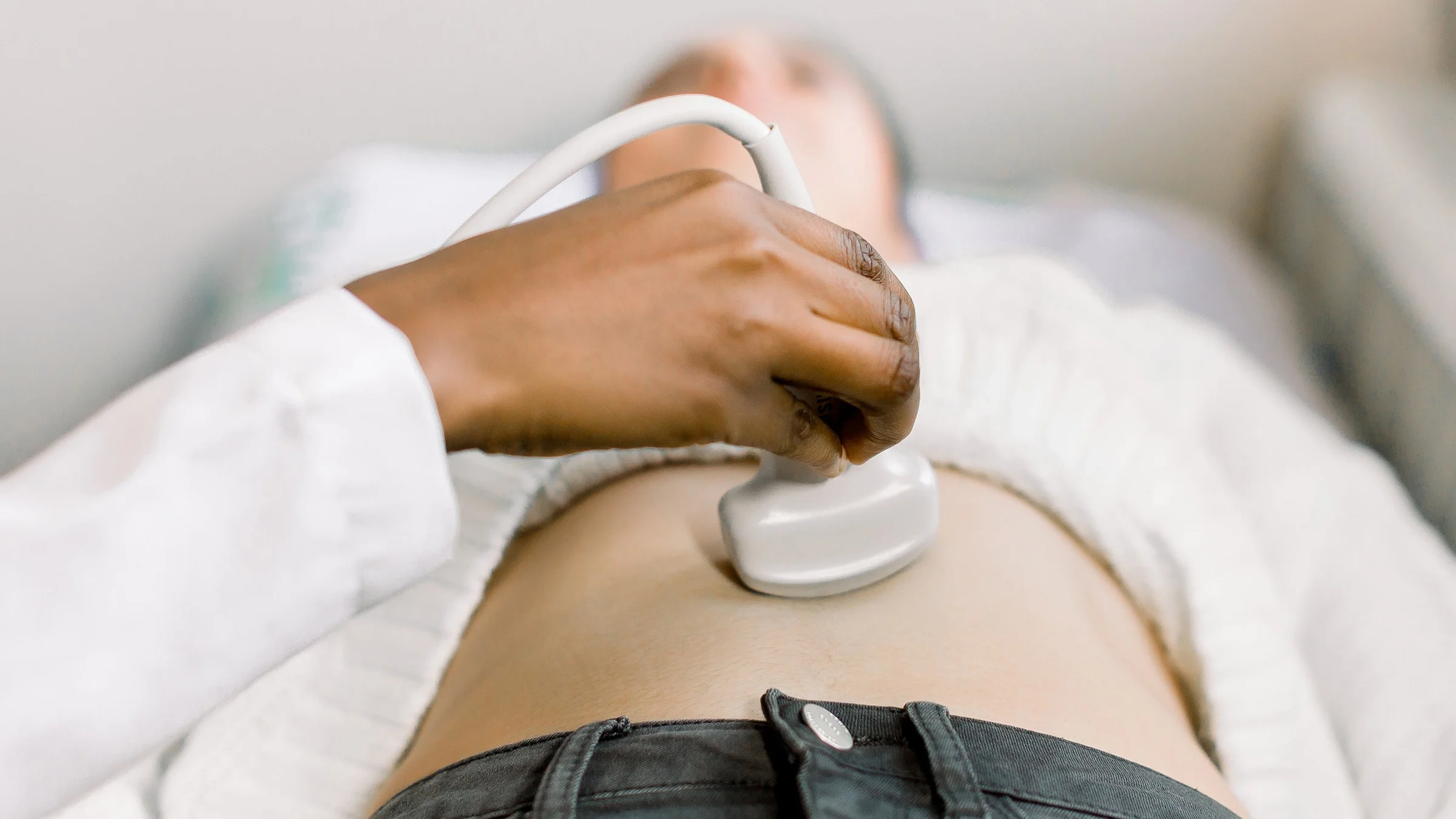 Close-up cropped shot of a person receiving an ultrasound scan. The focus is on the doctor's hand and the ultrasound transducer (a wand type device).