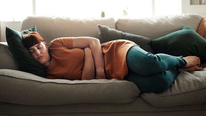 Young woman with bangs laying on the couch clutching her stomach in pain.