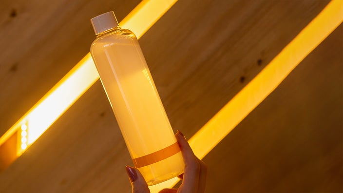 Bottom of shampoo being held up by a hand with nail polish as the light comes through the wooden background.