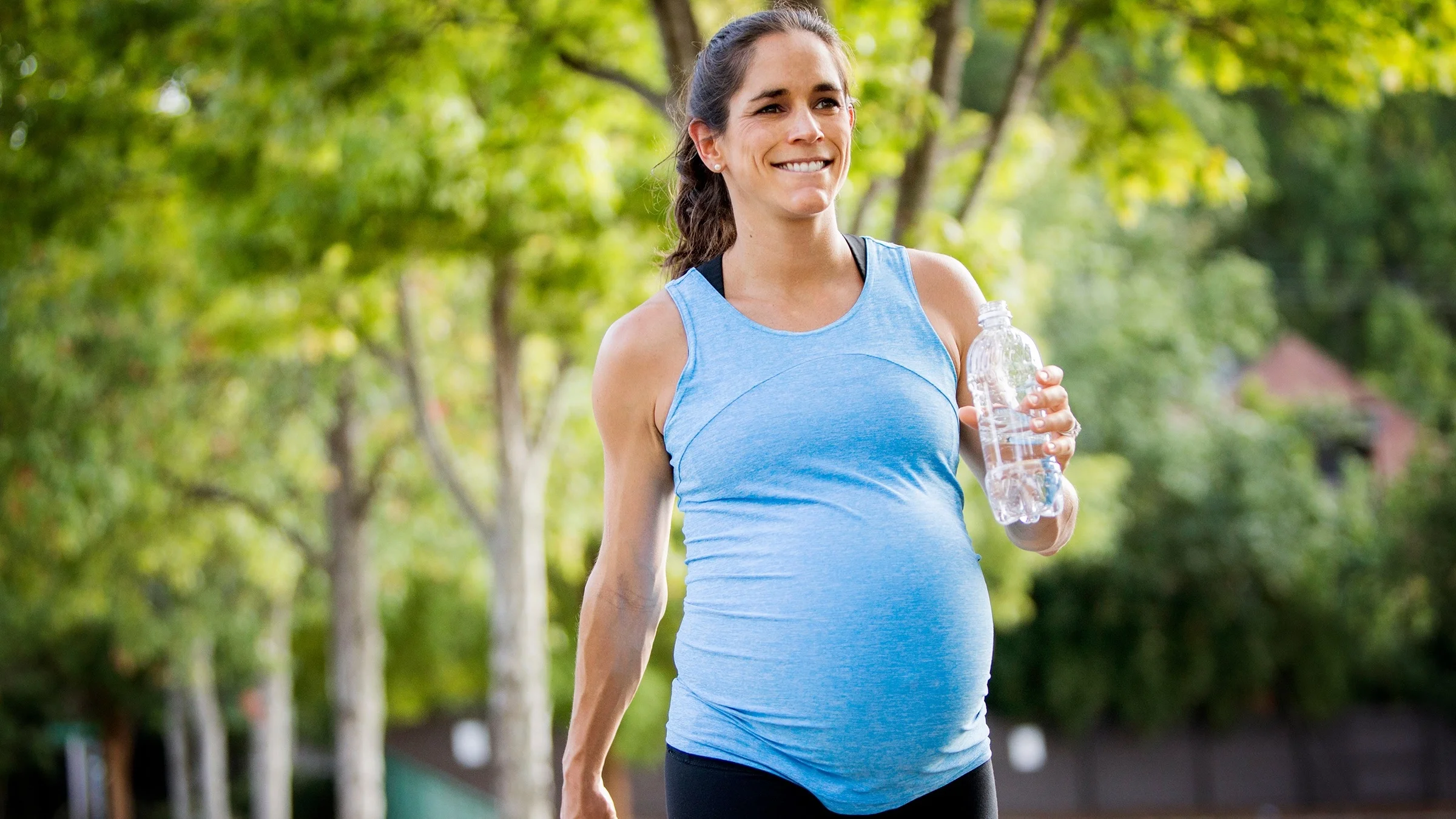A portrait of an extremely strong and muscular pregnant woman on a walk through the park. She is carrying a water bottle and smiling big.