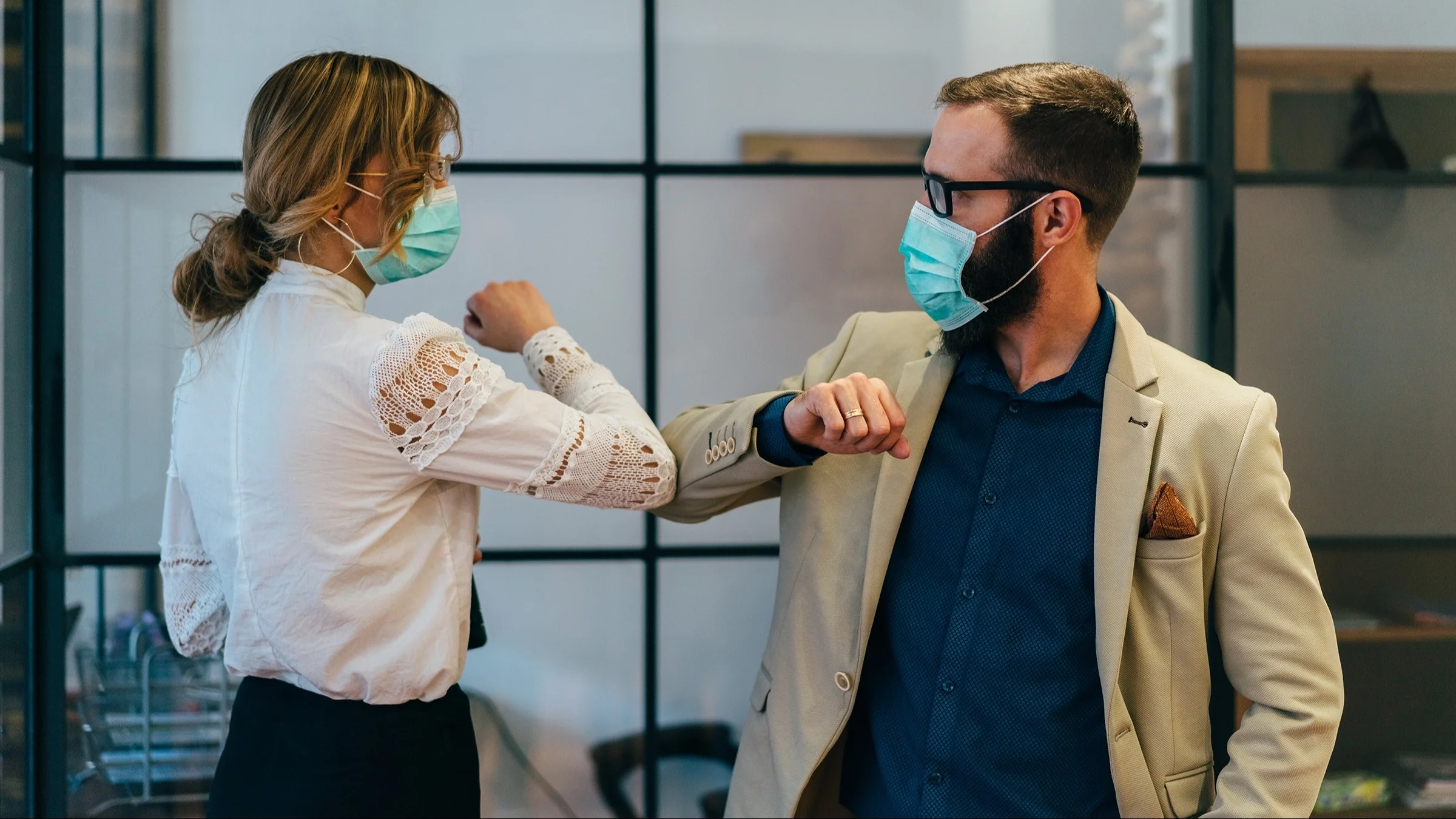 Two coworkers passing each other in the hallway touching elbows to say hello instead of shaking hands. They are both in professional corporate work clothes and blue medical face masks.