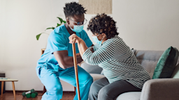A nurse helping a woman up off the couch in her home. She is using a wooden cane to push herself up as well.
Adene Sanchez/E+ via Getty Images