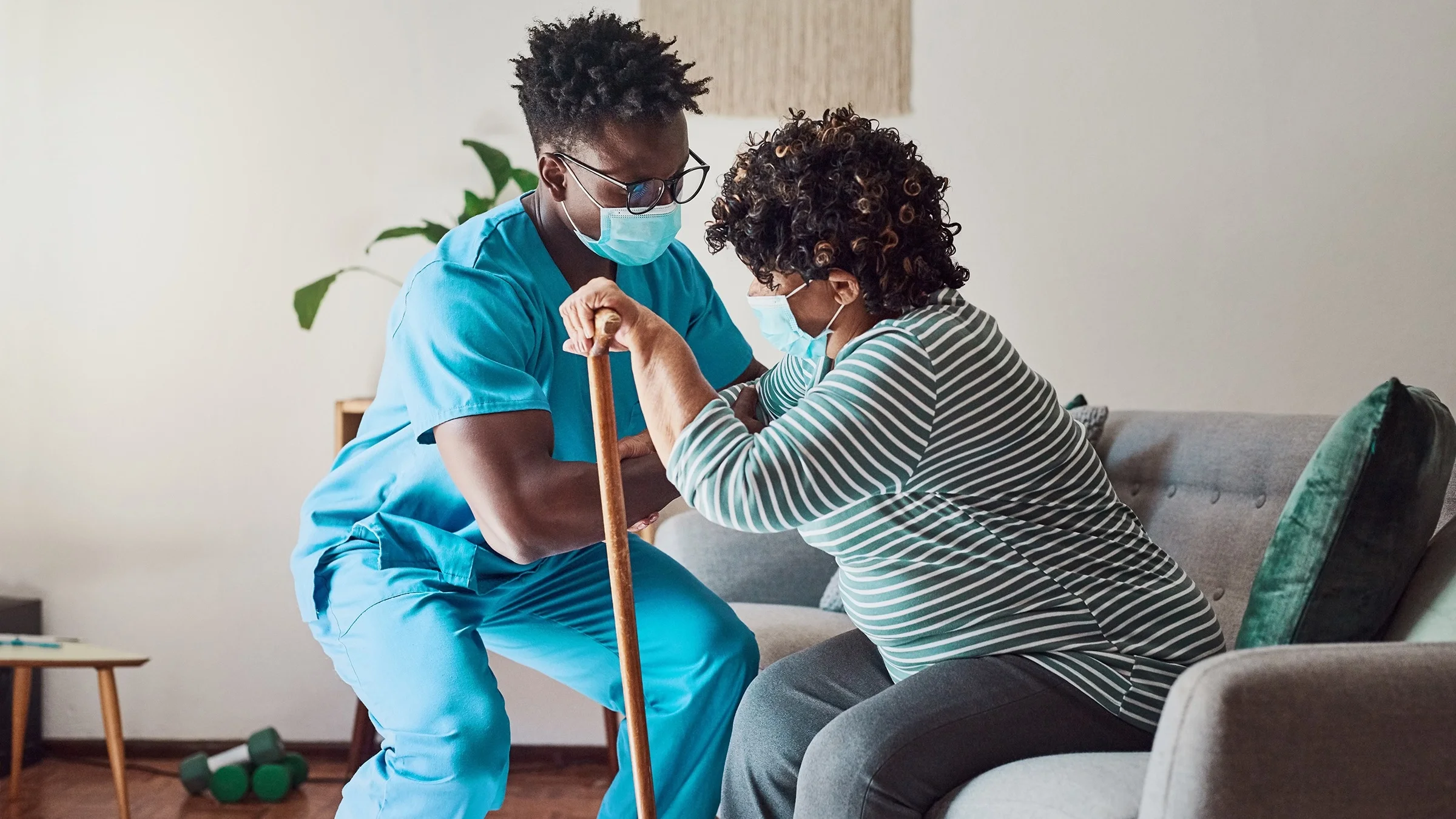 A nurse helping a woman up off the couch in her home. She is using a wooden cane to push herself up as well.