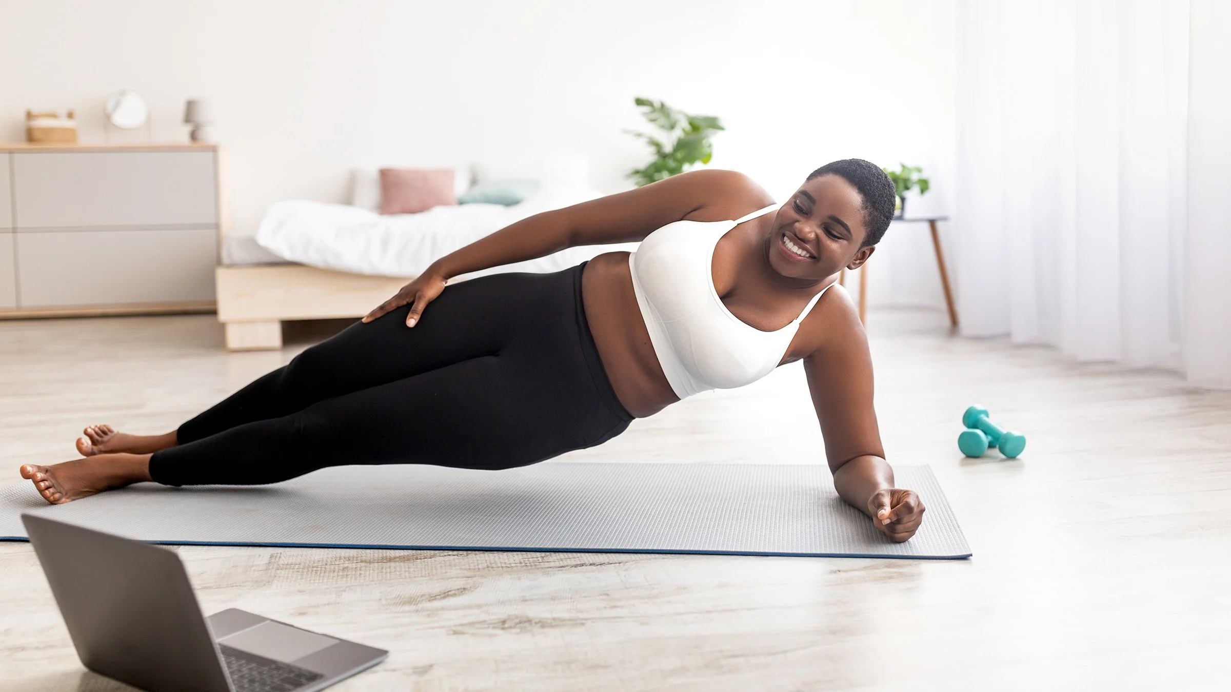 Young Black woman doing exercises in her living room. She is watching a lesson on her laptop while she does a side plank.