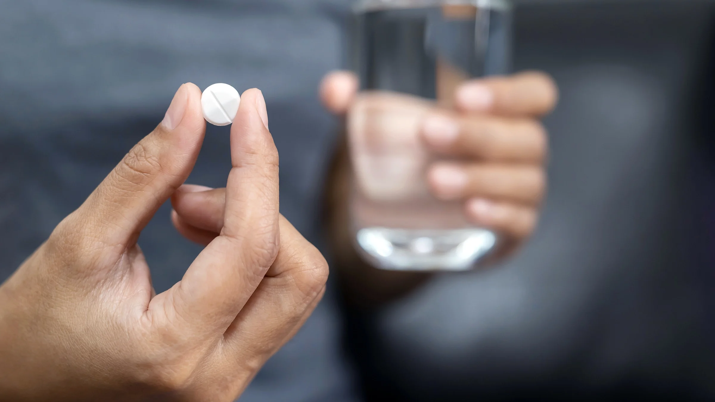 A person clutches a pill in between their thumb and forefinger in one hand and holds a glass of water in the other.
