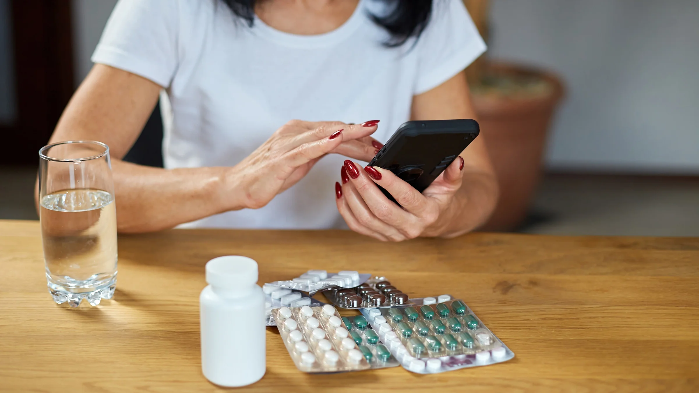 Cropped shot of woman reviewing medications on her phone. There are multiple blister packs of medication in front of her on the table.