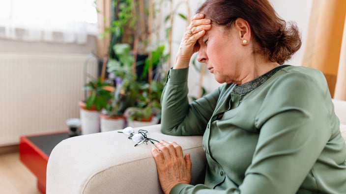 Elderly woman sitting on her couch resting her head in her hand in pain.