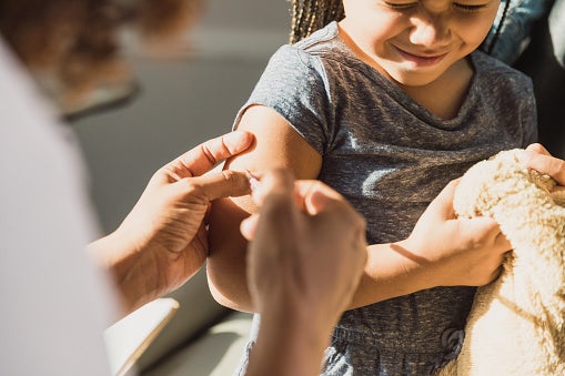 Close-up of a little girl getting a shot at the doctors office, holding her teddy bear and squinting her eyes.