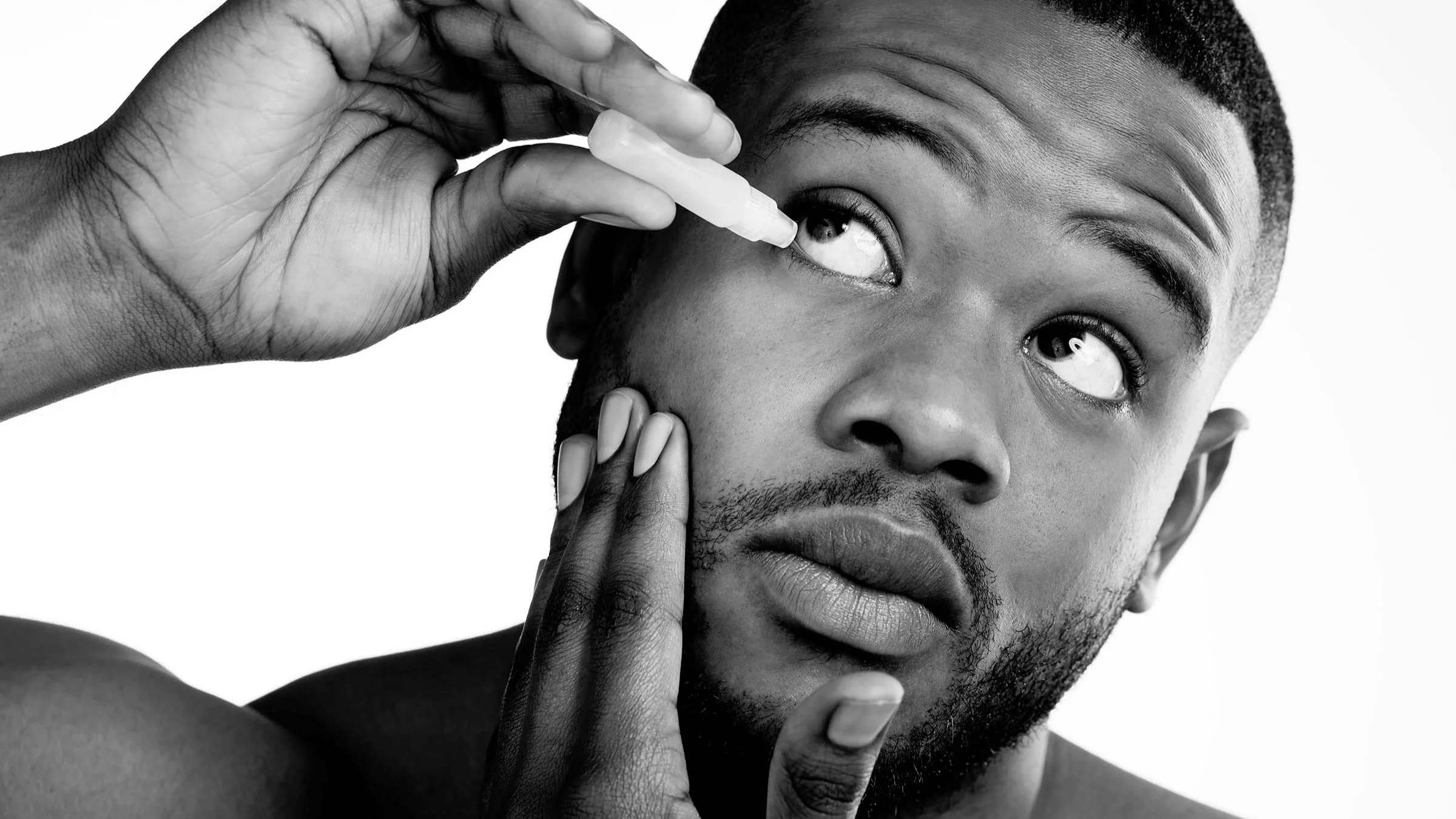 Black and white close-up of a man putting eye drops in his right eye on a white background.