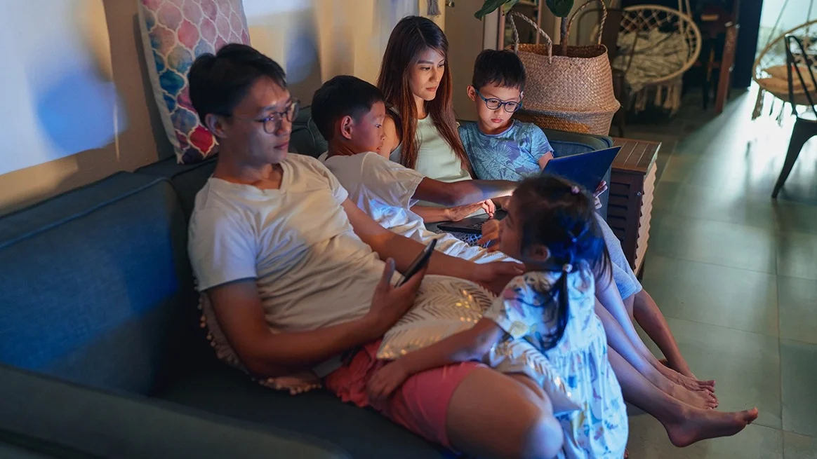 Entire family with two kids all on different electronic devices as they sit on their couch in a dimly lit room.