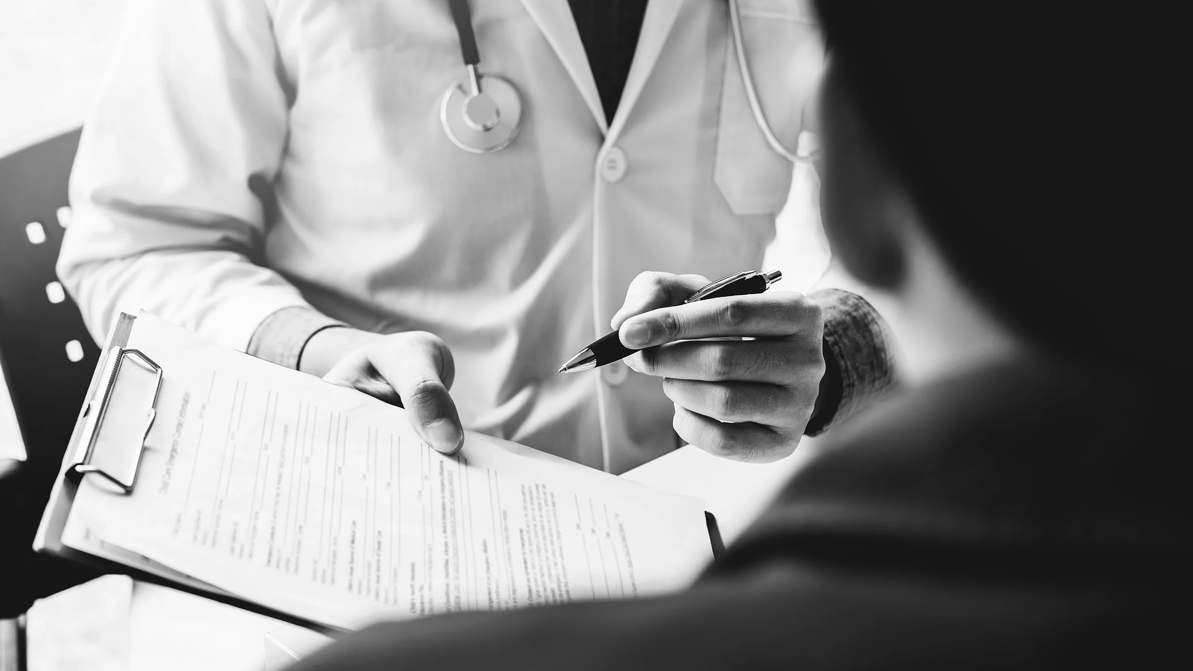 Black and white close-up of a doctor presenting medical paperwork to patient on clipboard.