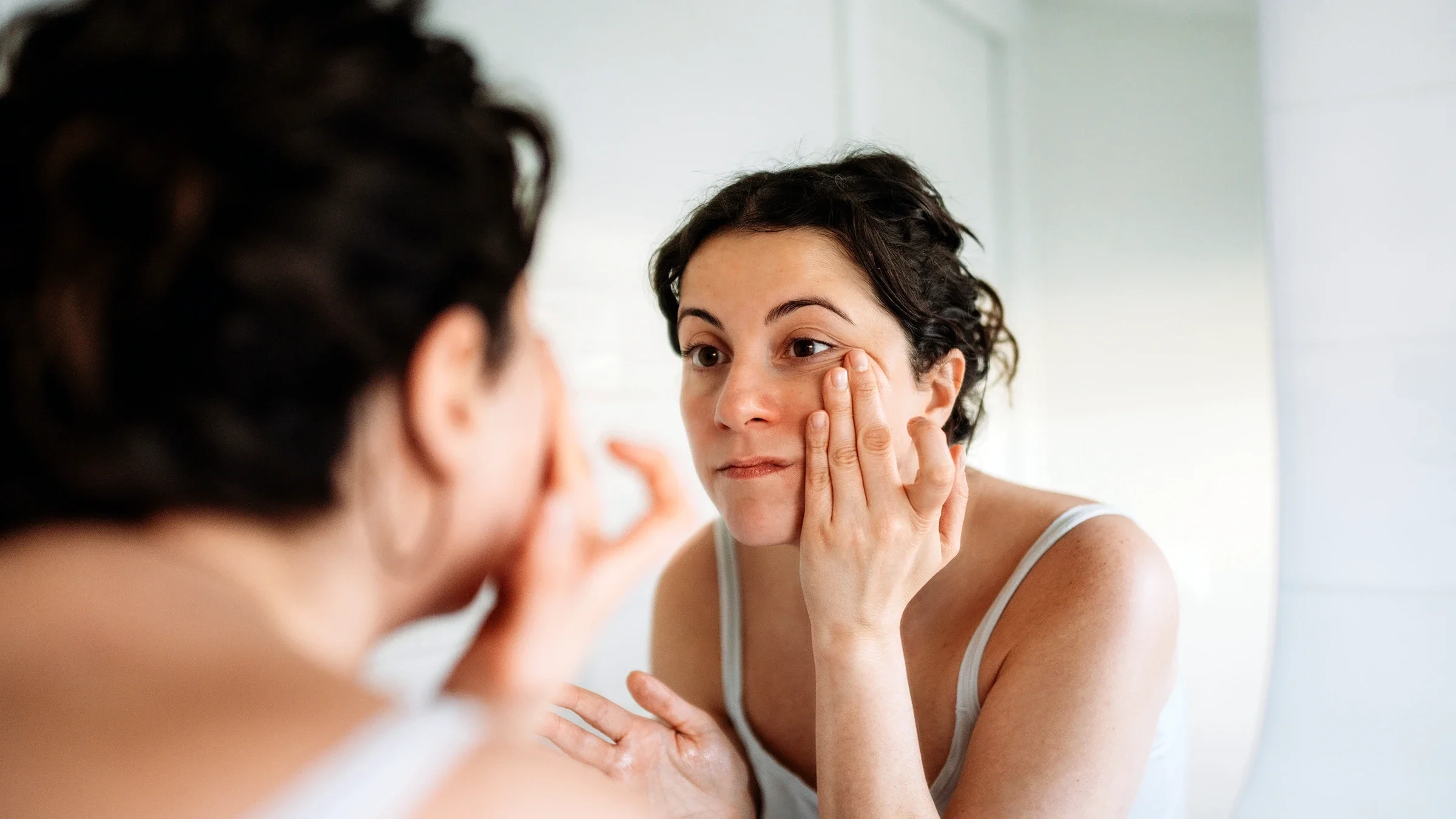 A woman touches her face as she looks at her reflection in the mirror.