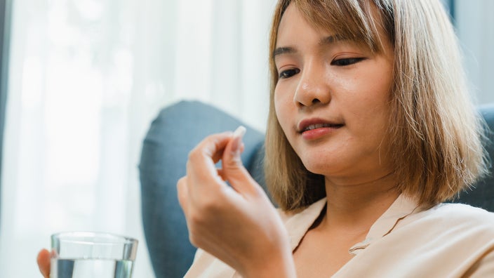 Woman holding a pill and a glass of water while sitting on the sofa at home.