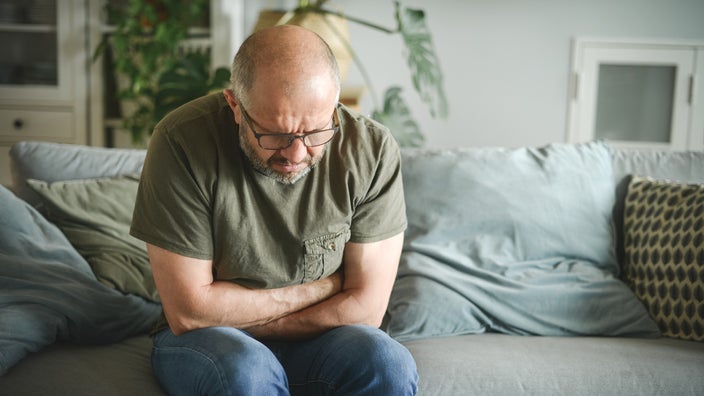 Older man with balding gray hair grasping his stomach in pain on the couch. He is wearing a green t-shirt with a pocket, jeans, and glasses.