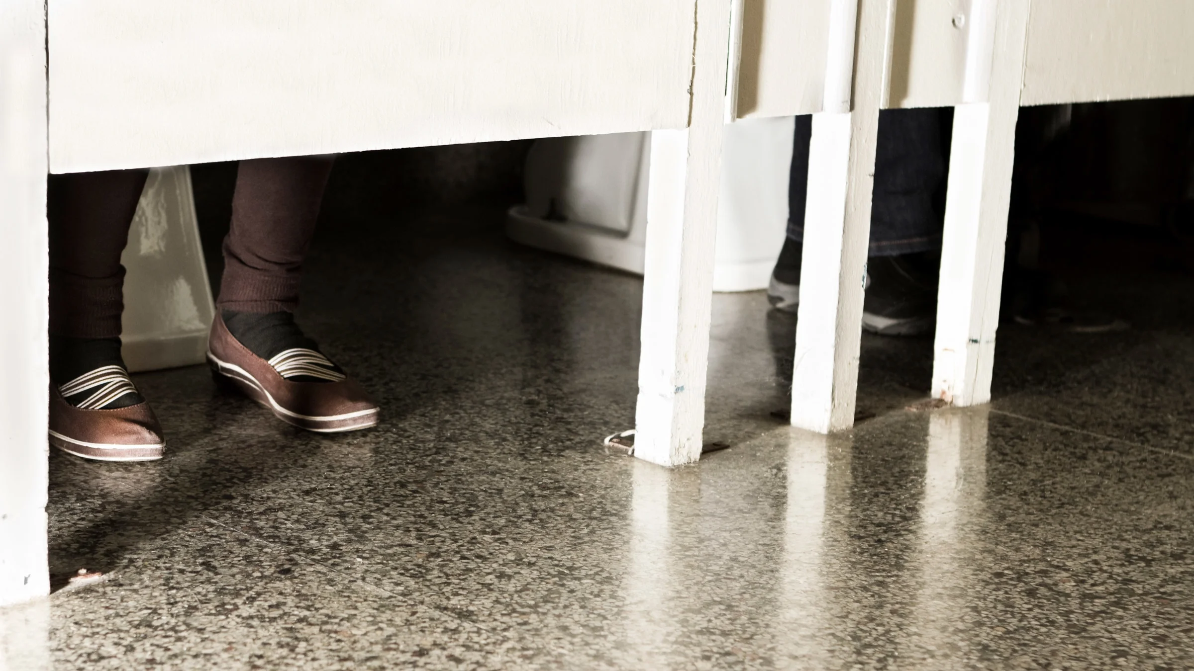 Shoes peeping out of two occupied bathroom stalls.