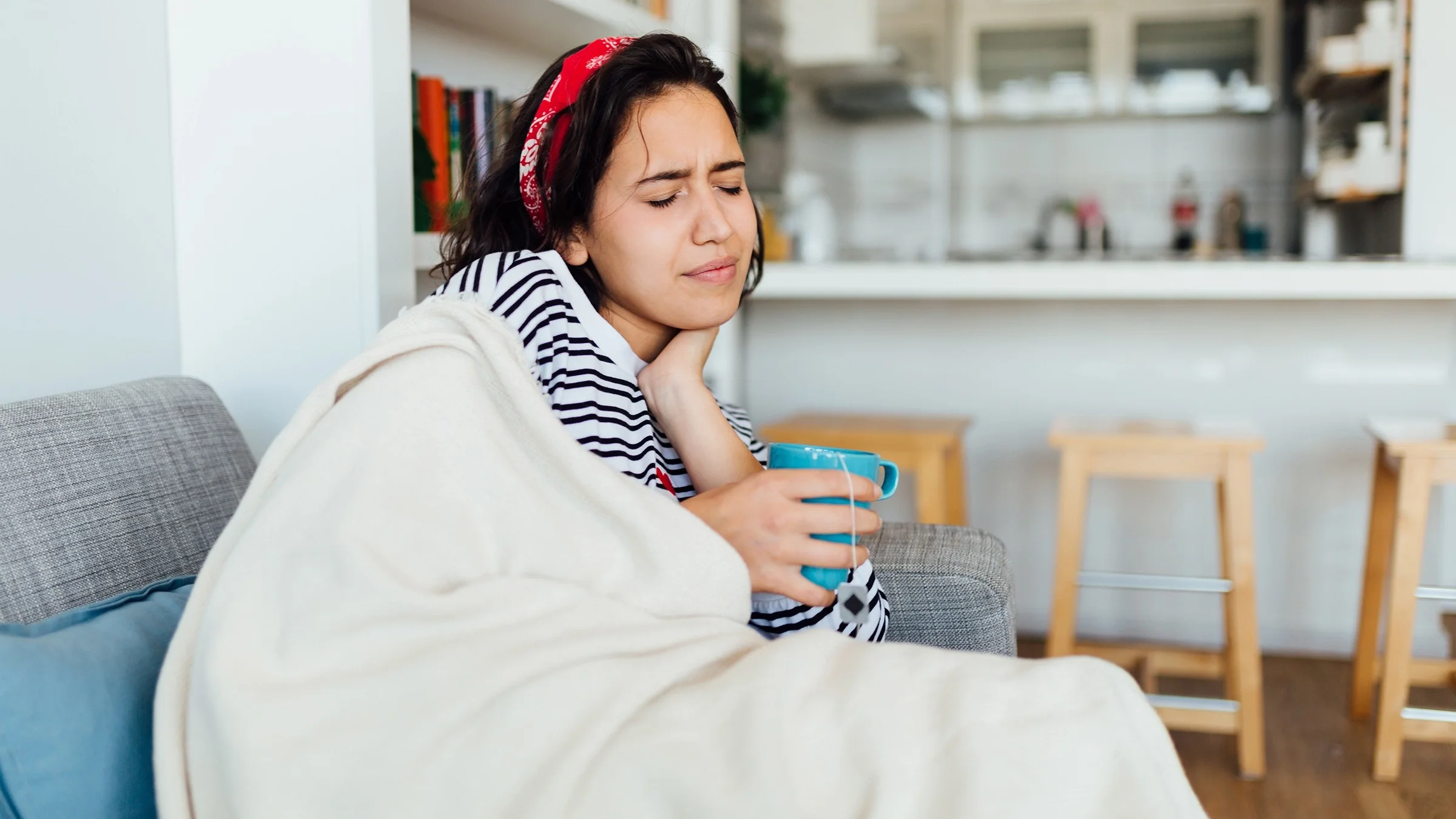 Young woman sitting on the couch with a cup of tea and holding her neck and throat.