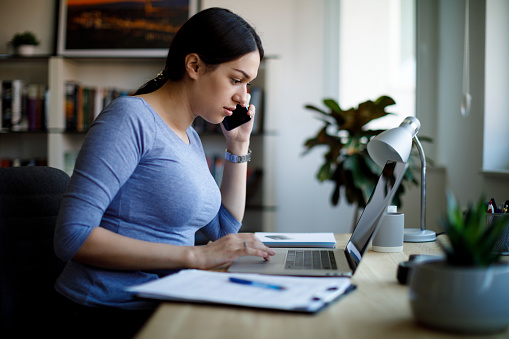 Woman talking on her phone while looking something up on her laptop at her at home desk.
