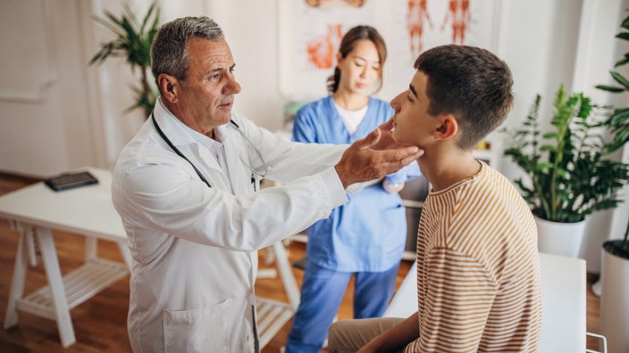 Young teenage boy at the doctor's office getting his lymph nodes examined. The nurse is in the background taking notes.