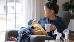 Woman holding her newborn and a coffee cup. There are breast pumps on the side table and she is looking out the window with a slightly sad expression.
FatCamera/E+ via Getty Images