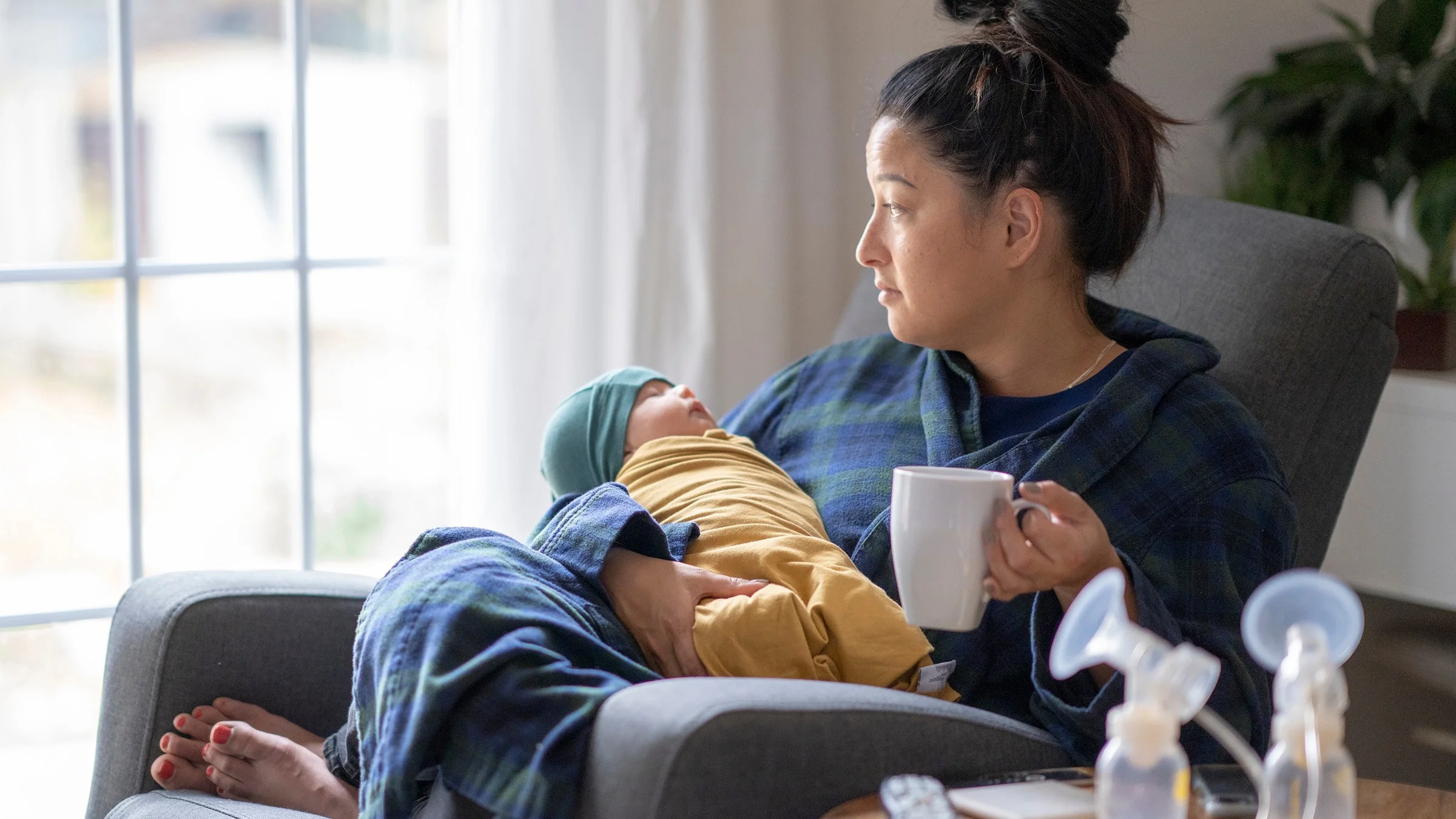 Woman holding her newborn and a coffee cup. There are breast pumps on the side table and she is looking out the window with a slightly sad expression.