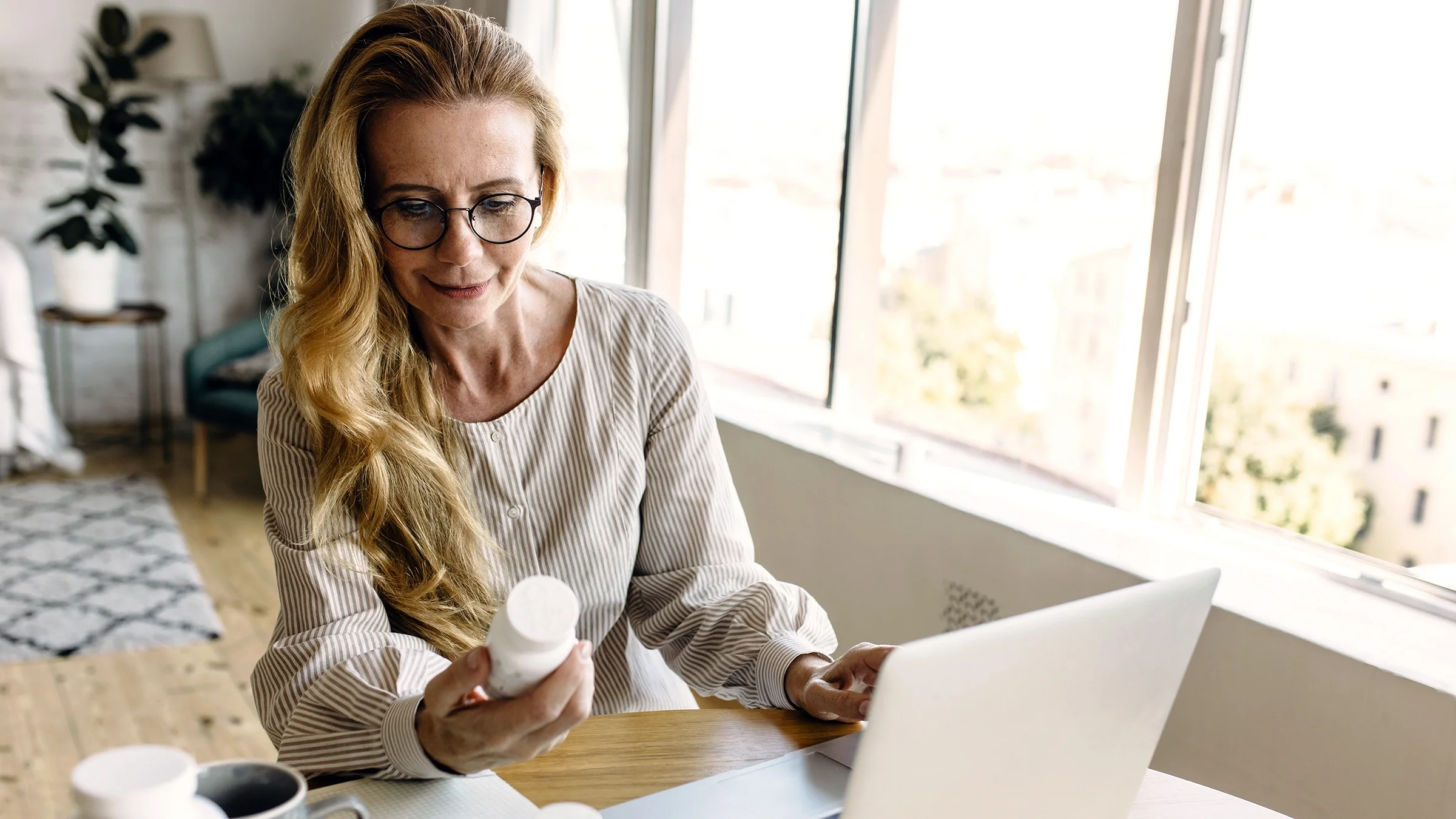 A woman sits at a table with a laptop while holding a prescription bottle.